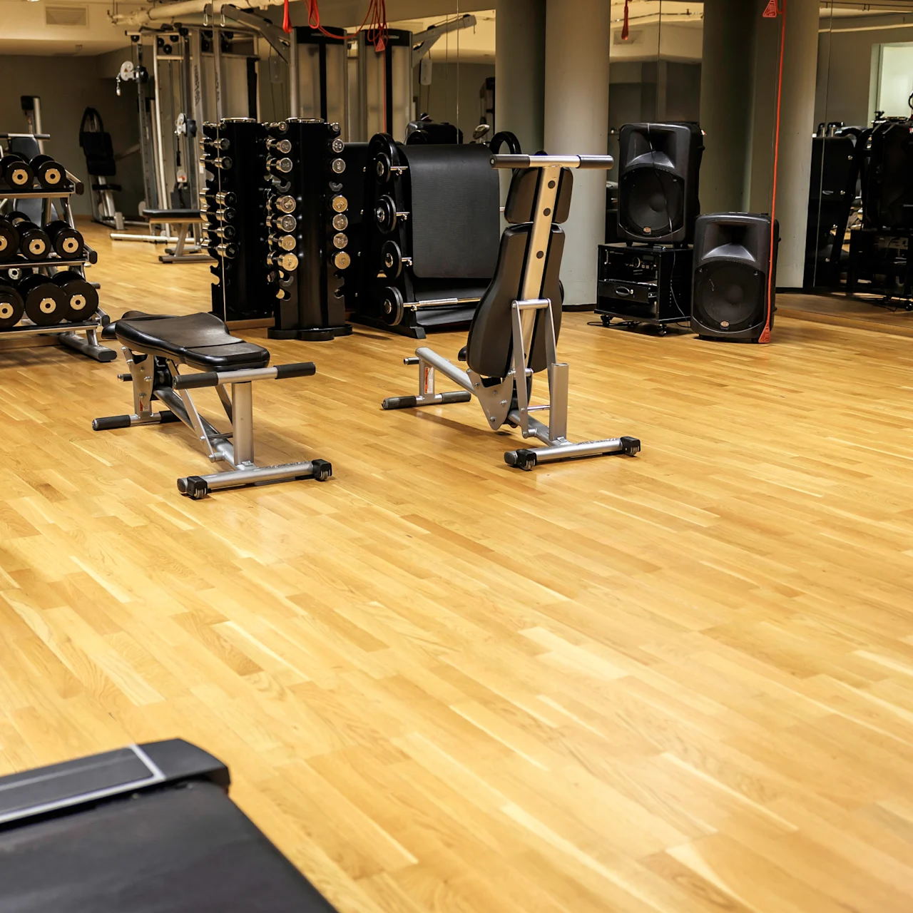 Gym equipment, including benches and weights, stands idle on a spacious wooden floor; mirrors and pillars surround, enhancing the room's open atmosphere.