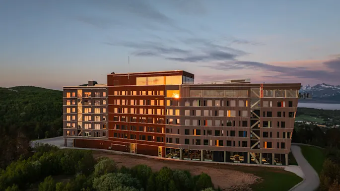 A modern multi-story wooden building reflects the sunset, with mountains and a fjord in the background. It stands surrounded by lush greenery.