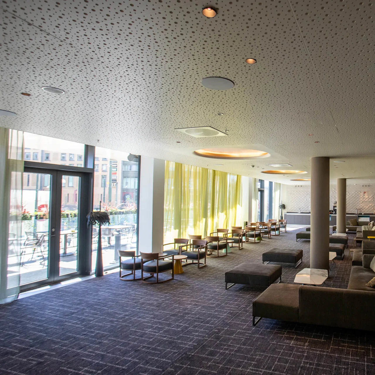 Lounge area with soft seating and chairs, arranged near large windows. Light streams through sheer curtains, illuminating a spacious, carpeted room. Modern ceiling design with circular lights and columns.