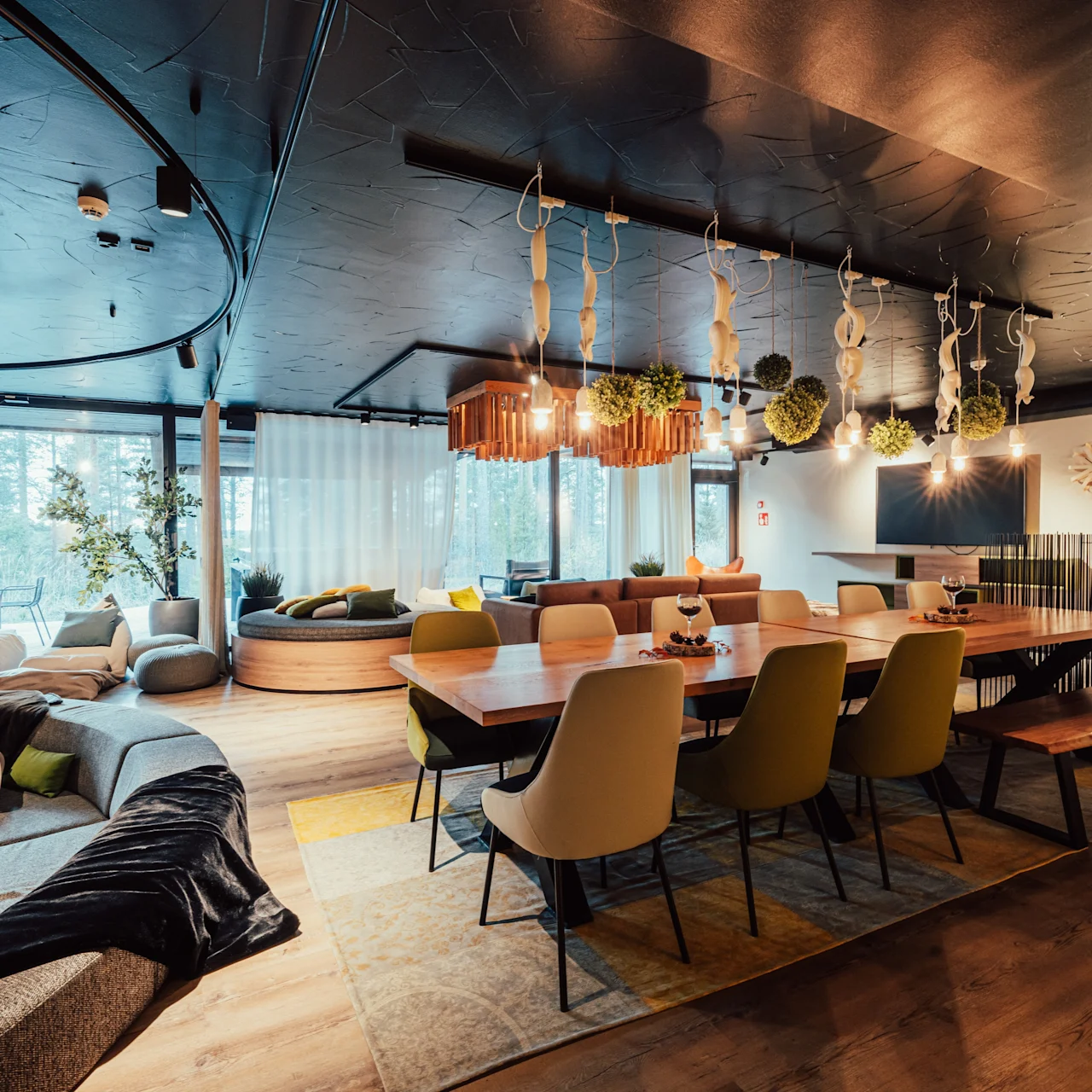 Spacious lounge features a large wooden dining table with chairs, surrounded by modern couches and greenery hanging from the ceiling. Floor-to-ceiling windows provide natural light.