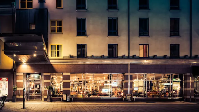 En bygningfasade, opplyst om natten, viser store vinduer som avslører et varmt opplyst interiør med mennesker inni. Et skilt med "Göpik" er synlig til venstre.