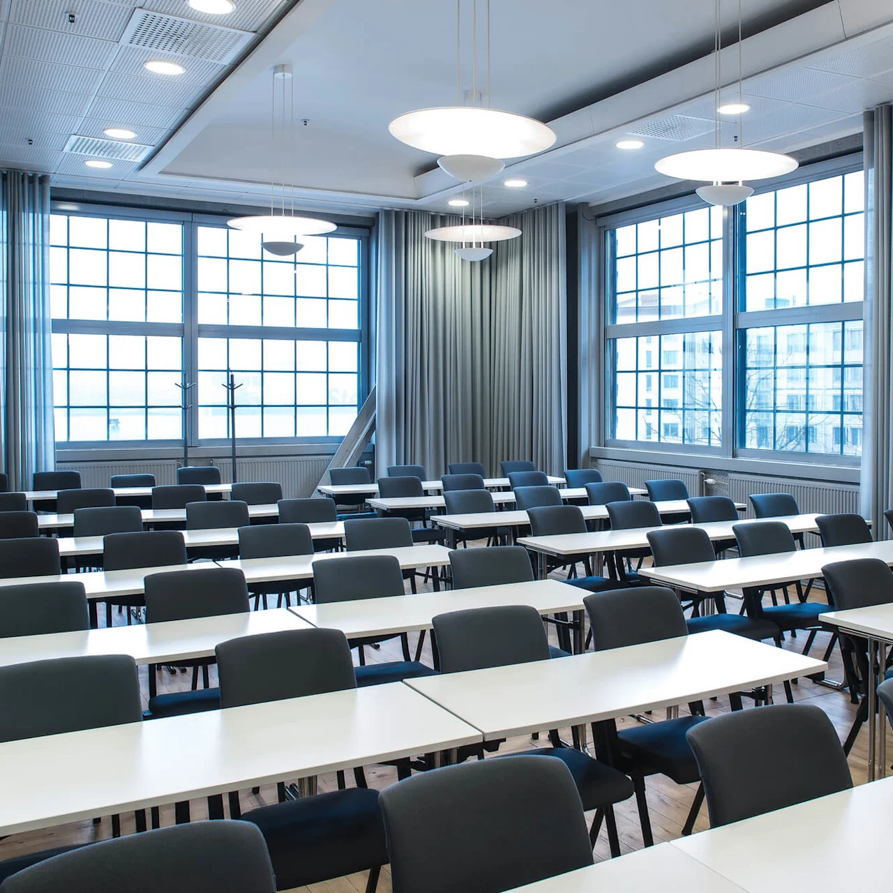 Rows of empty tables and chairs are arranged in a bright classroom. Large windows with curtains allow natural light, while overhead lights illuminate the space.