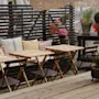 Träbord och stolar är arrangerade på en uteplats, tillsammans med färgglada kuddar, krukväxter och ett trägaller, vilket skapar en mysig och inbjudande atmosfär.