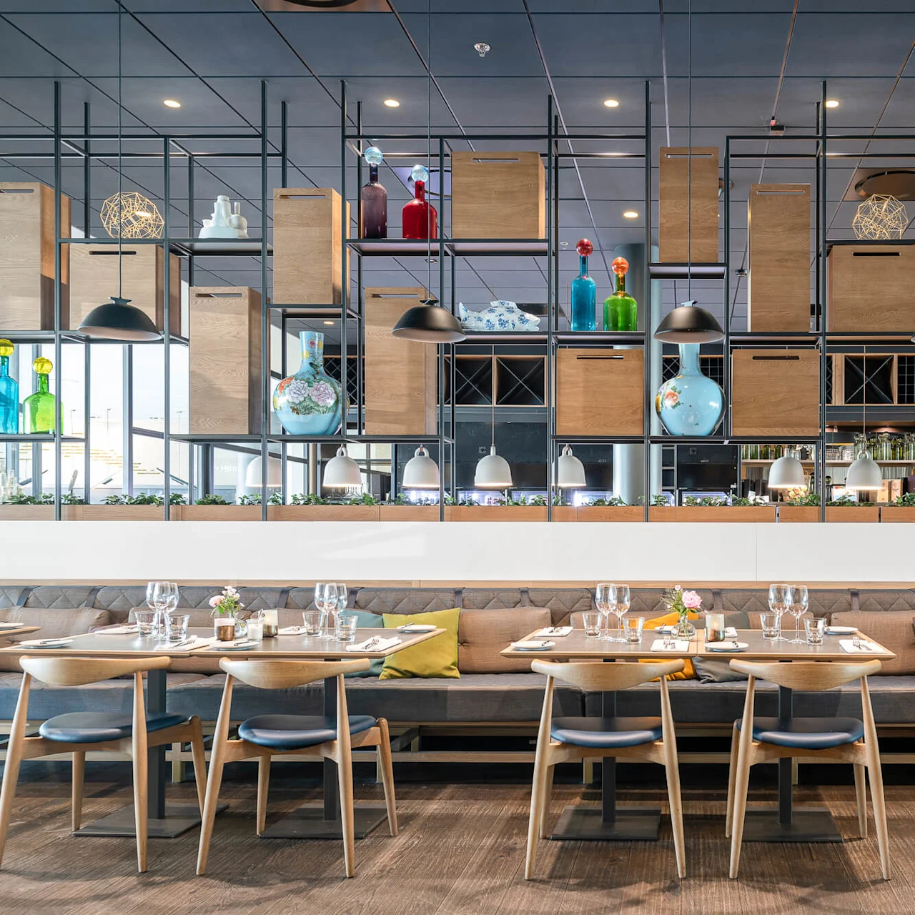 Wood dining tables set with glassware, napkins, and flowers are arranged in a modern restaurant with artistic shelving and colorful vases, under a ceiling with hanging lights.