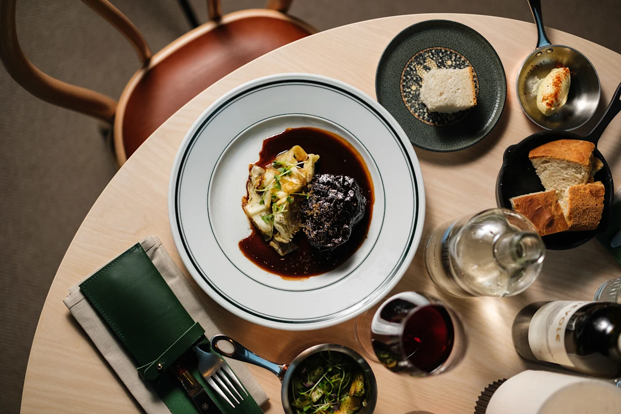 Ett bord med en huvudrätt bestående av kött och grönsaker, omgiven av bröd, smör, grönsaker, ett glas rödvin och en vattenflaska i en matsituation.