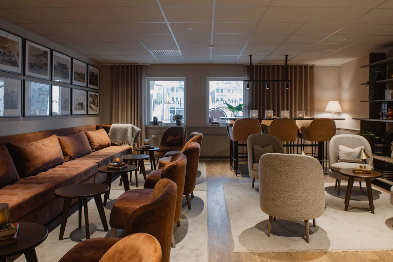 Et varmt og indbydende loungeområde med komfortable siddepladser, herunder en lang sofa og lænestole, samt en bardisk. Synlig tekst: FREDAGATEN.