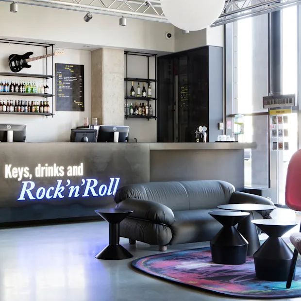 A modern reception desk displays the text "Keys, drinks and Rock'n'Roll" with a guitar and bottles above. Comfortable seating and colorful decor enhance the welcoming lounge atmosphere.