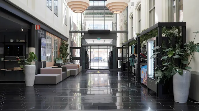 An indoor lobby with sofas, potted plants, and a glass entrance door, reflecting a glossy tiled floor.