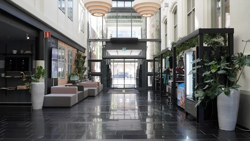 An indoor lobby with sofas, potted plants, and a glass entrance door, reflecting a glossy tiled floor.