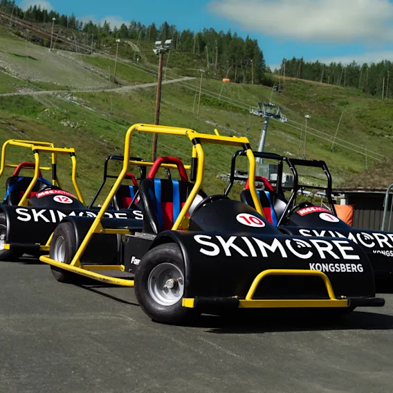Tre svarte downhill-karter med gule veltbøyler og hvit "Skimore Kongsberg"-tekst parkert på asfalt. I bakgrunnen ses en grønn skibakke med en skiheis om sommeren under en blå himmel.