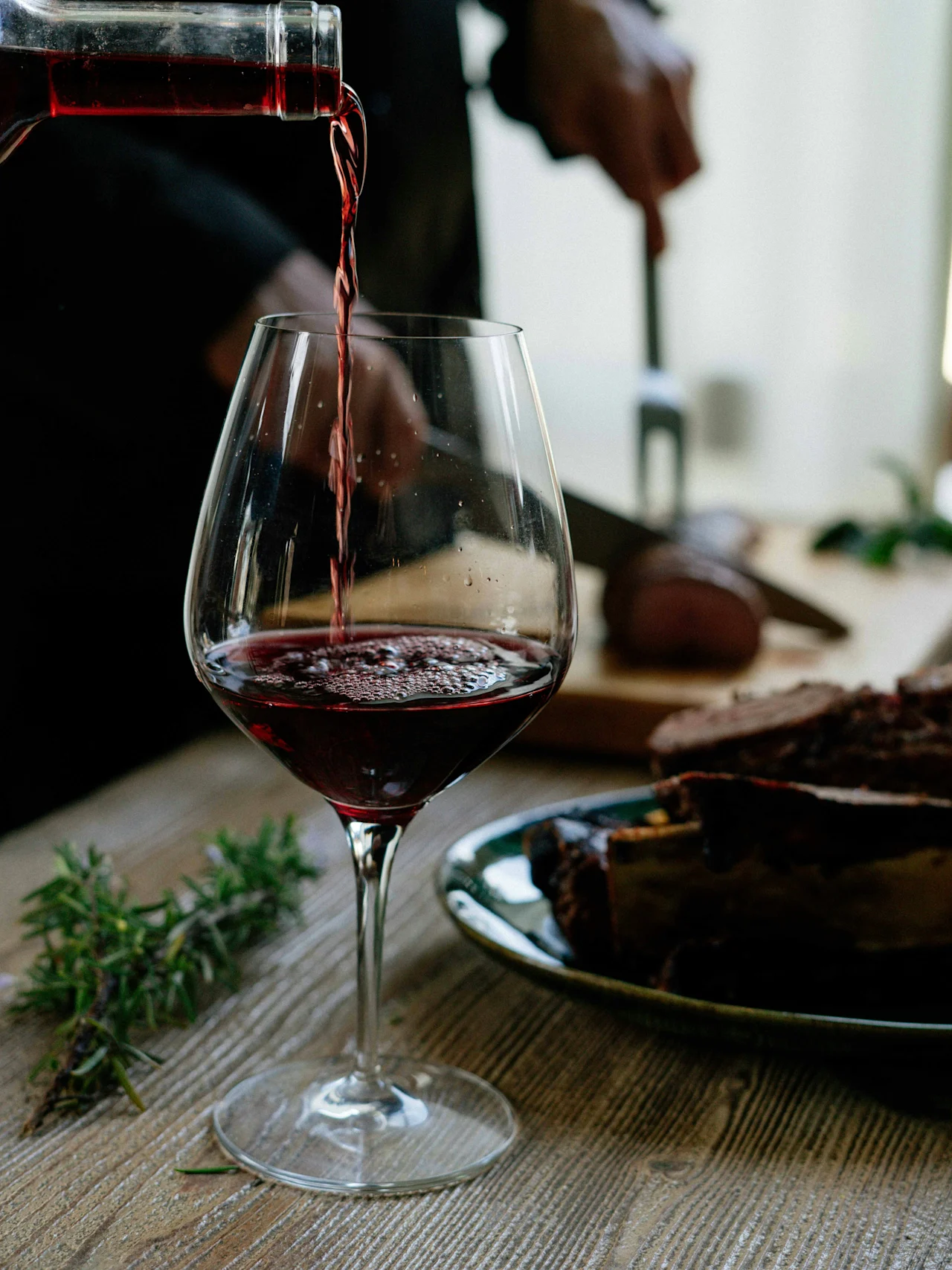 Red wine pours into a glass, while in the background, a person slices roast meat on a wooden cutting board beside a sprig of rosemary.