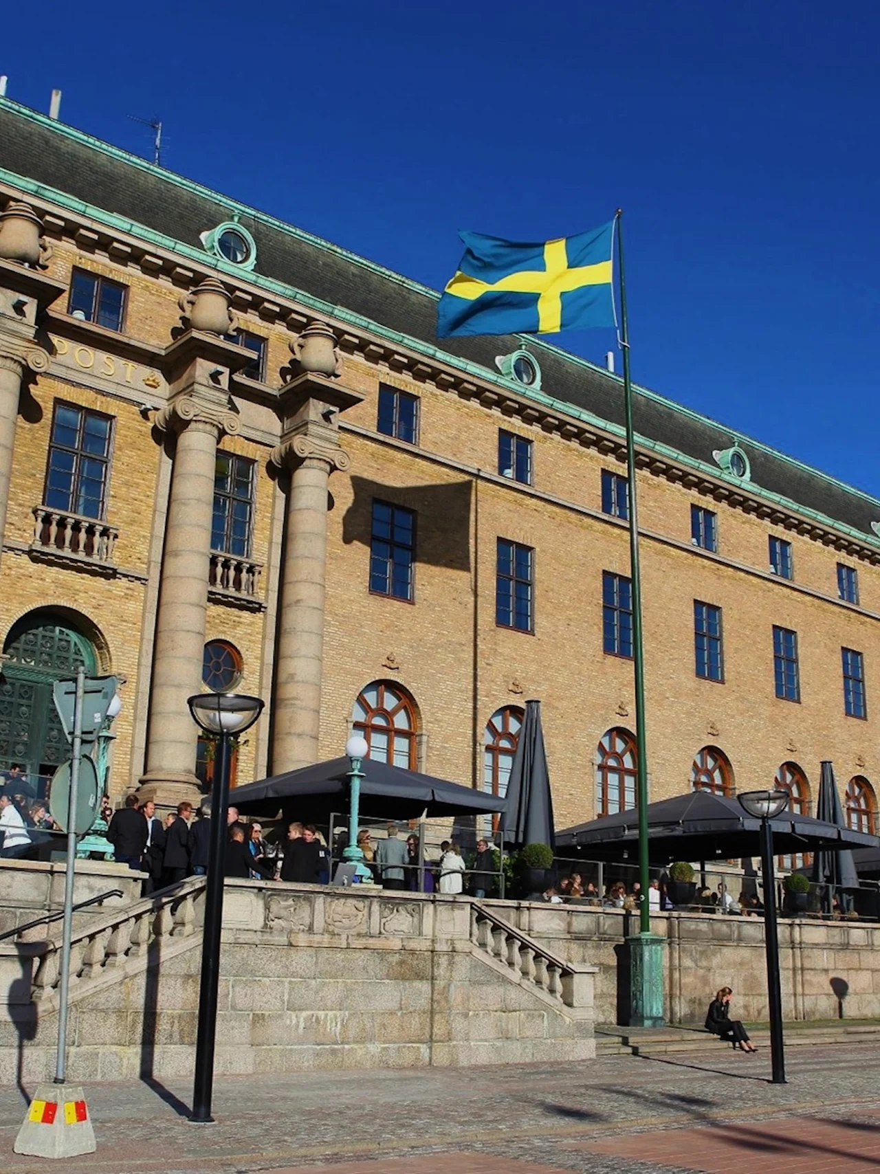 En ståtlig byggnad med texten POST, en svensk flagga vajar och människor njuter av ett utomhuskafé en solig dag.