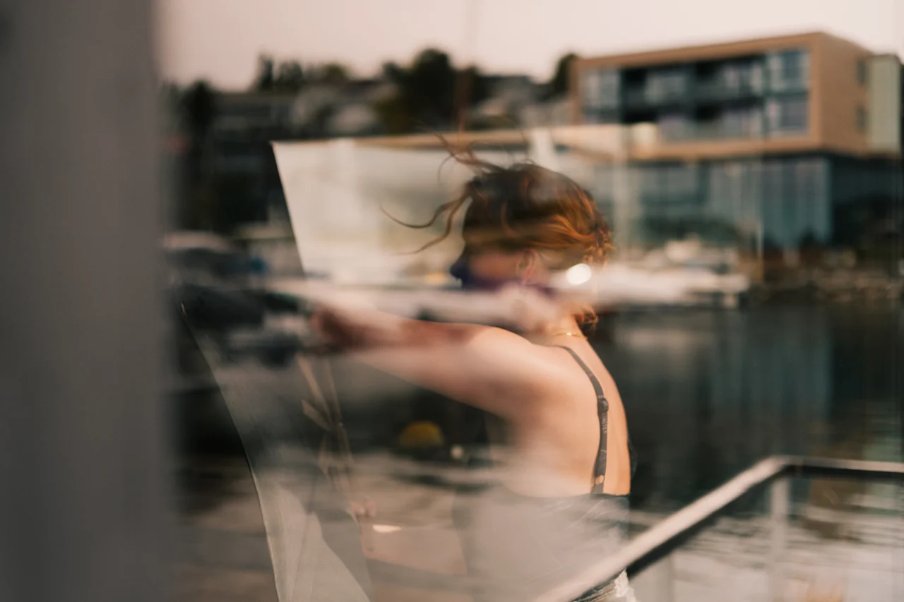 En person maler udendørs ved vandet, med bygninger og både i baggrunden. Håret blæser let i vinden.