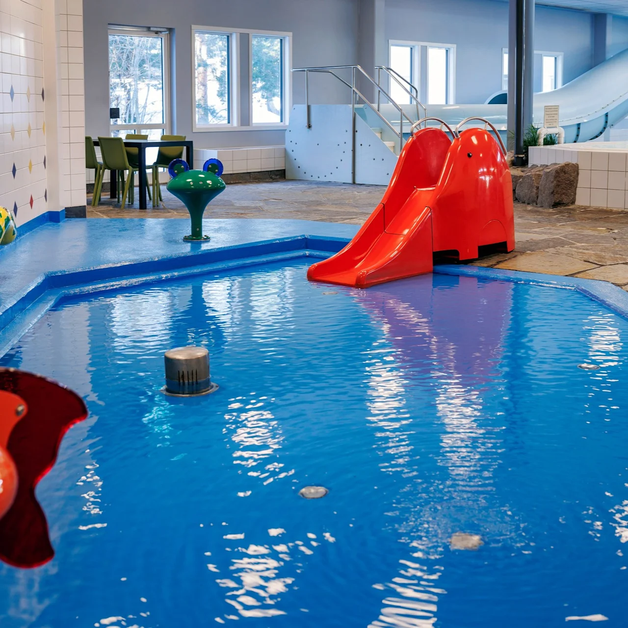 Children's pool area featuring a red slide entering shallow water, surrounded by cartoon animal figures and playful designs, with windows and tables in the background.