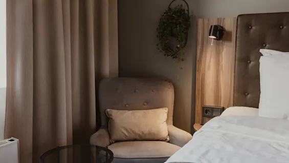 Viihtyisä hotellihuoneen nurkkaus, jossa on mukava nojatuoli, sänky valkoisilla liinavaatteilla ja lämmin, kutsuva tunnelma.