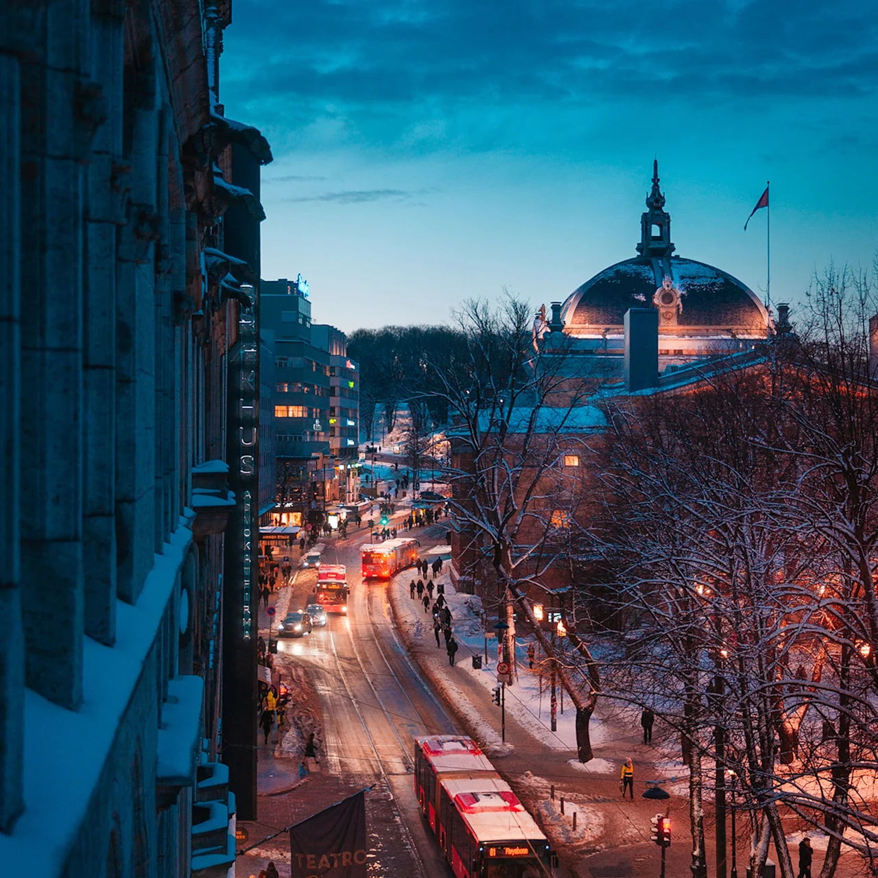 Buses drive on a snow-covered city street at dusk, surrounded by illuminated buildings and leafless trees. Signs read "TEATRO" and "HOTEL CHRISTIANIA TEATER."