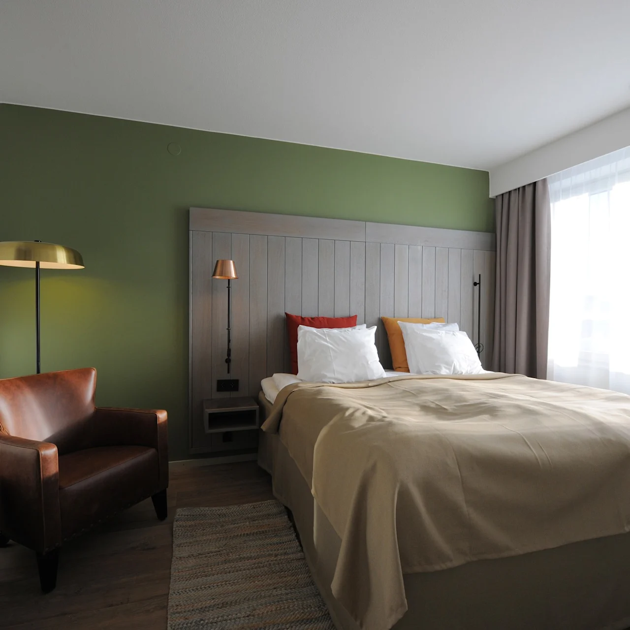 A neatly made bed with beige linens is set against a green accent wall. An armchair and floor lamp stand nearby. Sunlight filters through large curtains.