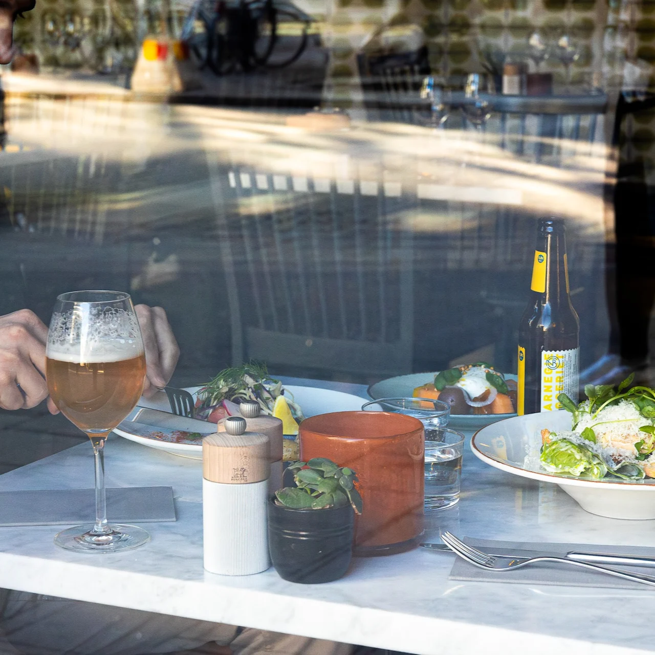 A person dines at a restaurant table, using utensils on a dish with vegetables, accompanied by a glass of beer, bottled drink, and potted plants, viewed through a window.