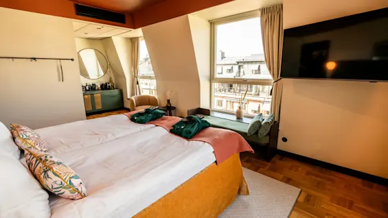 A neatly made bed with decorative pillows faces a television, surrounded by modern furnishings and large windows, in a cozy hotel room. Bathrobes are placed at the bed's foot.