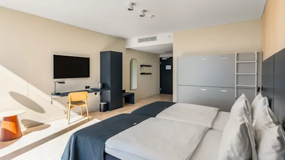 Et lyst og moderne hotelværelse med to senge, et skrivebordsområde og en vægmonteret køjeseng, badet i naturligt lys.