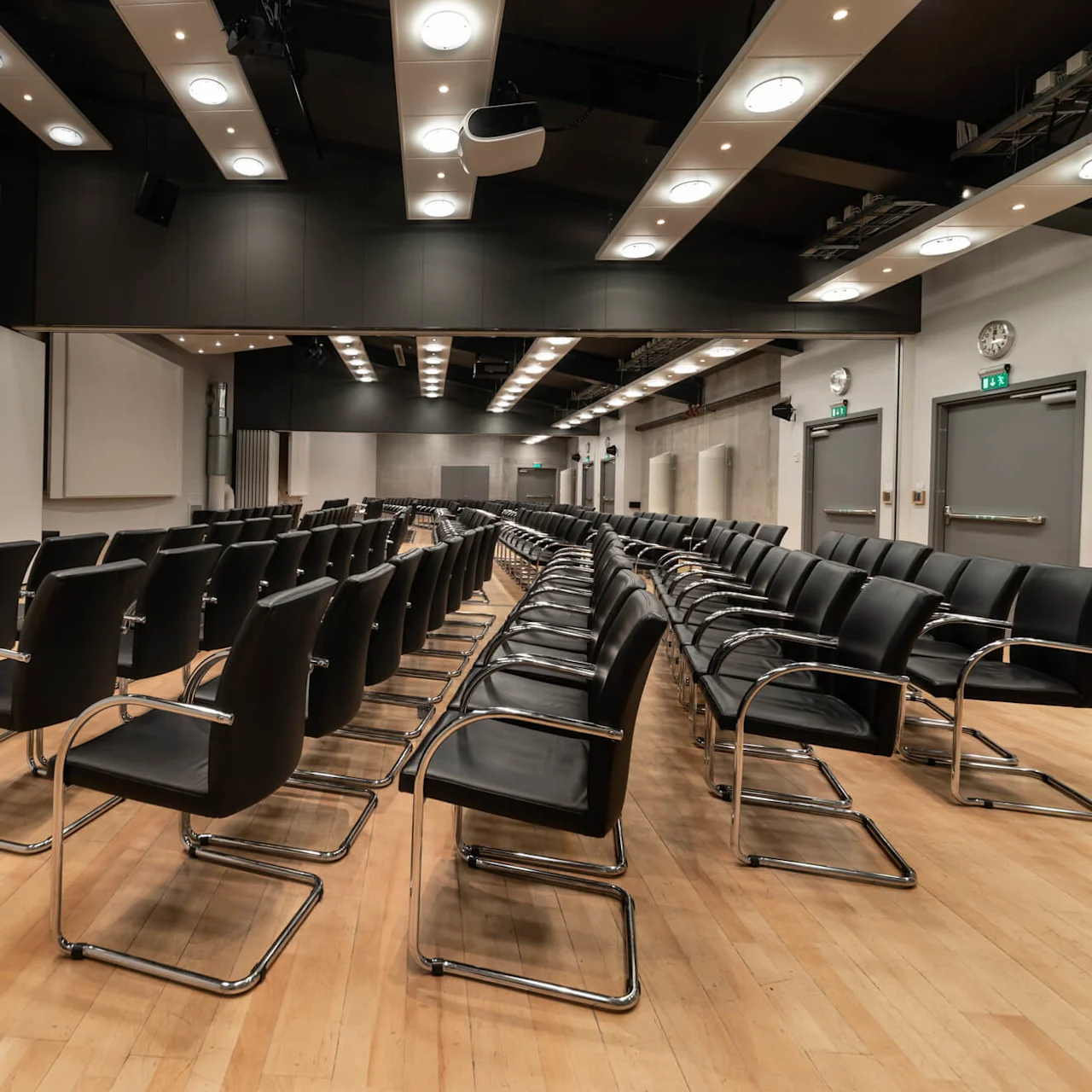 Flera rader med svarta stolar är prydligt uppradade i ett rymligt konferensrum med trägolv och stora projektionsskärmar.