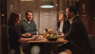 Fire venner som ler og prater rundt et middagsbord i et koselig og varmt opplyst restaurantlokale.