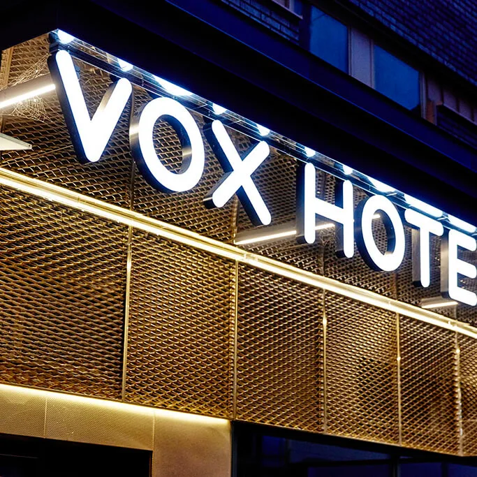 Et stort skilt viser "VOX HOTEL" i lyse, hvide bogstaver. Skiltet er monteret på en metallisk, netbaggrund, oplyst af lys i et bymiljø.