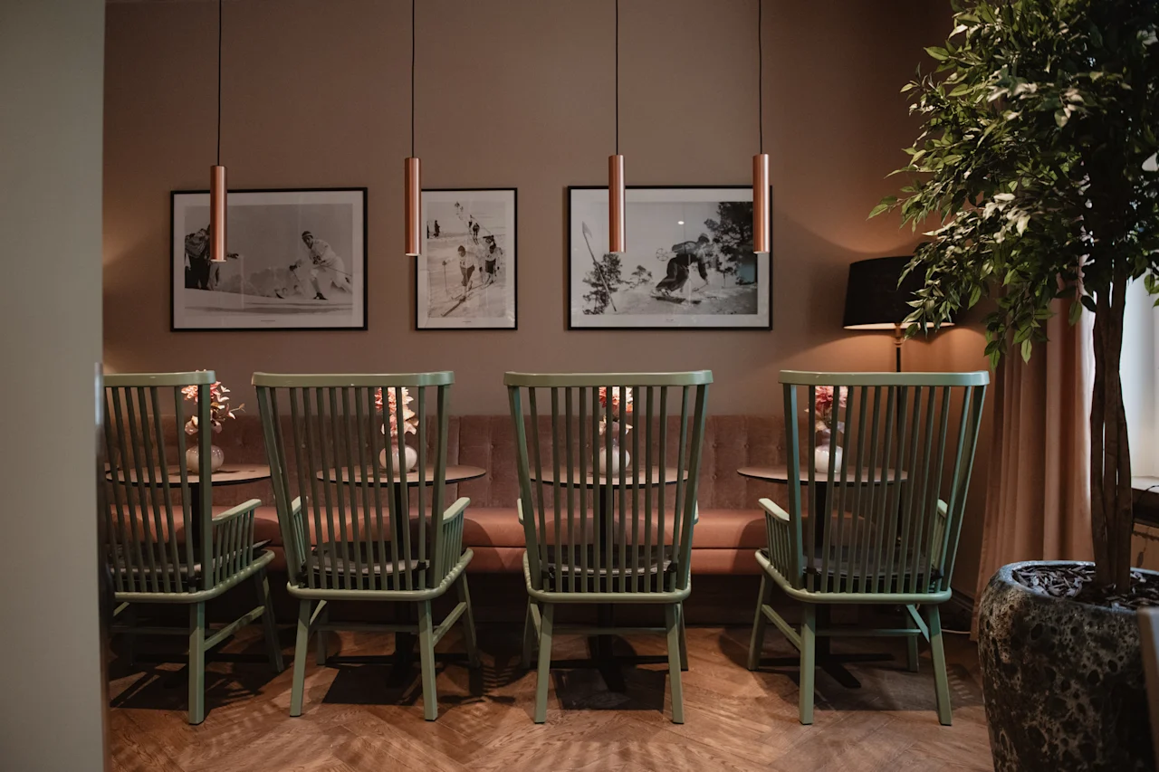 Et koselig og innbydende loungeområde med grønne trestoler, små bord, en komfortabel brun sofa og innrammede kunstverk.