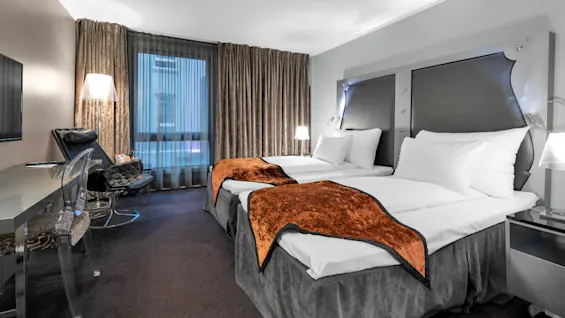 To pænt redte senge med orange tæpper står i et moderne hotelværelse. Et skrivebord, stol og lampe er i nærheden, med gardiner der dækker et vindue.