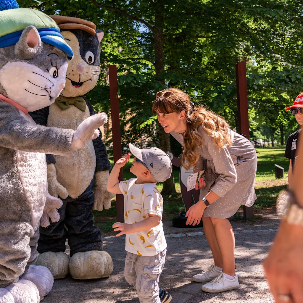 Barn och vuxna interagerar med kattmaskotar i en park, möjligen i Uppsala Pelle Svanslösparken.