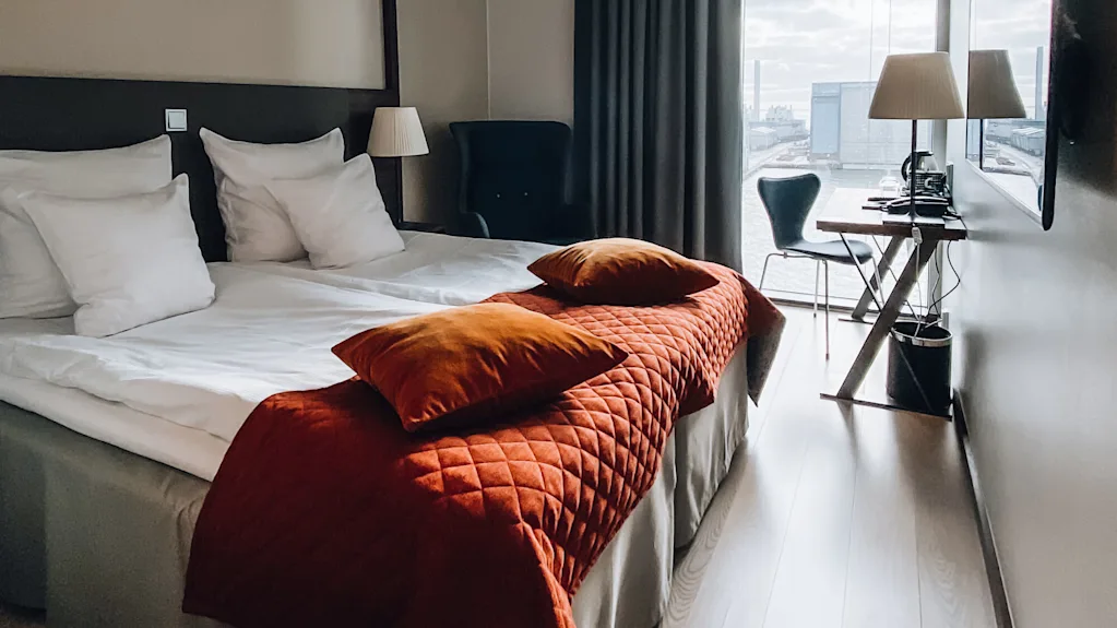 En prydligt bäddad säng med vita lakan och orange kuddar står i ett modernt hotellrum med ett minimalistiskt skrivbord och stol nära ett stort fönster med utsikt över en industriell strand.