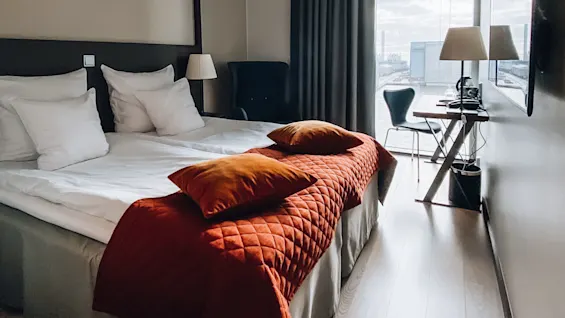 En pænt redt seng med hvidt sengetøj og orange puder står i et moderne hotelværelse med et minimalistisk skrivebord og stol nær et stort vindue med udsigt over en industriel havn.