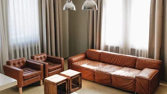 A brown leather sofa set sits in a sunlit room with beige curtains, metallic ceiling lamps, and wooden side tables beside a bed.