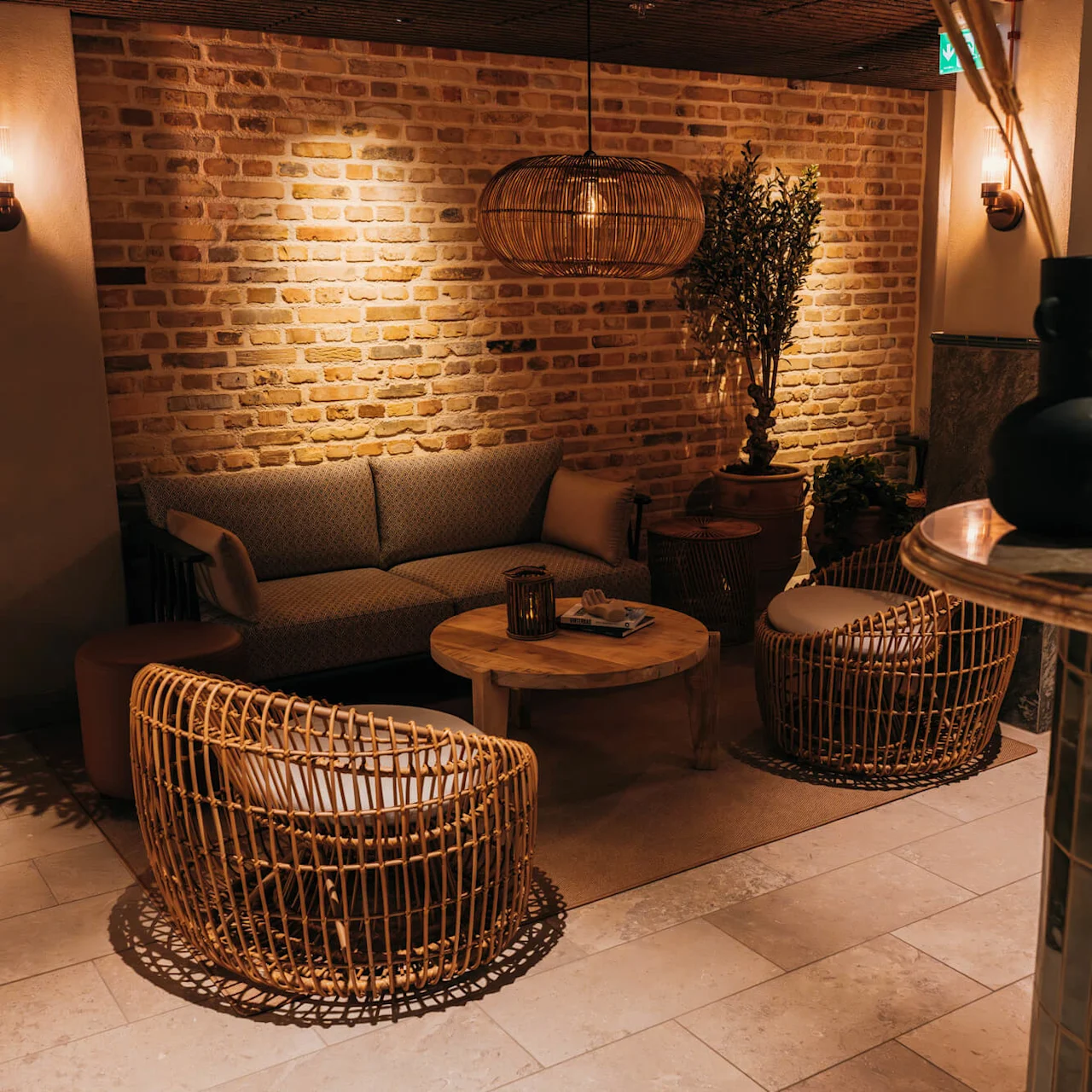 A cozy seating area with a sofa and wicker chairs surrounds a wooden coffee table. Ambient lighting and potted plants accent the brick wall in a warmly lit room.