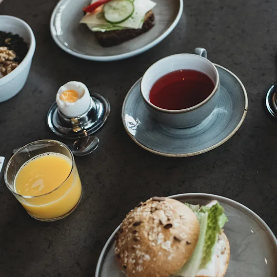 En frukostuppsättning med en smörgås, yoghurtskålar med nötter och bär, ett löskokt ägg, en kopp te och ett glas apelsinjuice på ett mörkt bord.