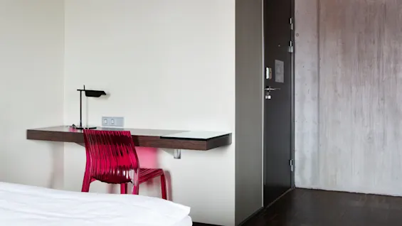 En rosa stol står ved et lite trebord med en svart lampe, ved siden av en lukket mørk dør i et minimalistisk rom med hvite og grå vegger.
