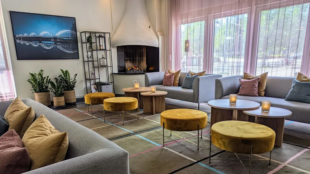 Soffor med färgglada kuddar omringar runda bord med ljus. Gula pallar står på en mönstrad matta. En öppen spis och väggkonst skapar en mysig atmosfär vid stora fönster.