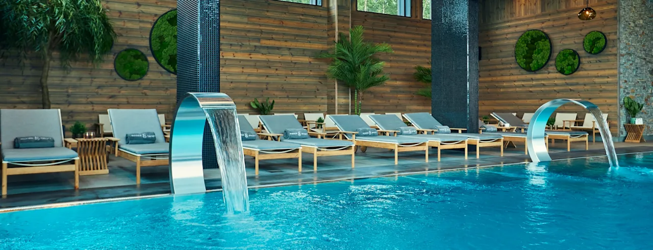A luxurious indoor pool features water cascading from steel fountains. Surrounding the pool are wooden lounge chairs with cushions, lush greenery, and large, decorative wall accents.