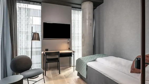 Ett modernt hotellrum har en enkelsäng med en grön filt, en väggmonterad TV ovanför ett skrivbord och en stor betongpelare. Naturligt ljus filtreras genom skira gardiner.