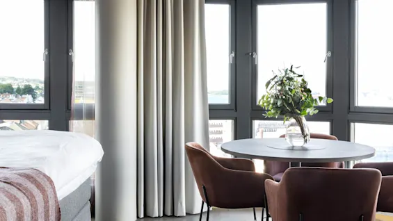 Bed with a striped blanket beside a pillar, next to large windows. Round table with brown chairs and a vase of flowers. Urban view outside.