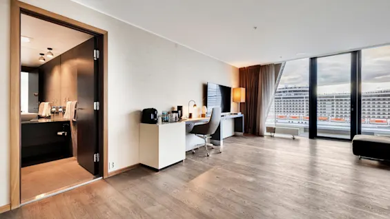 A modern hotel room features a workspace with a desk, chair, and large TV. Floor-to-ceiling windows offer a city view. An adjoining bathroom has towels and lighting fixtures.
