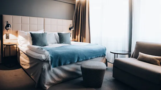 A neatly made bed with blue and white bedding is flanked by lamps in a softly lit modern bedroom with brown curtains, a cushioned headboard, a sofa, and a round ottoman.
