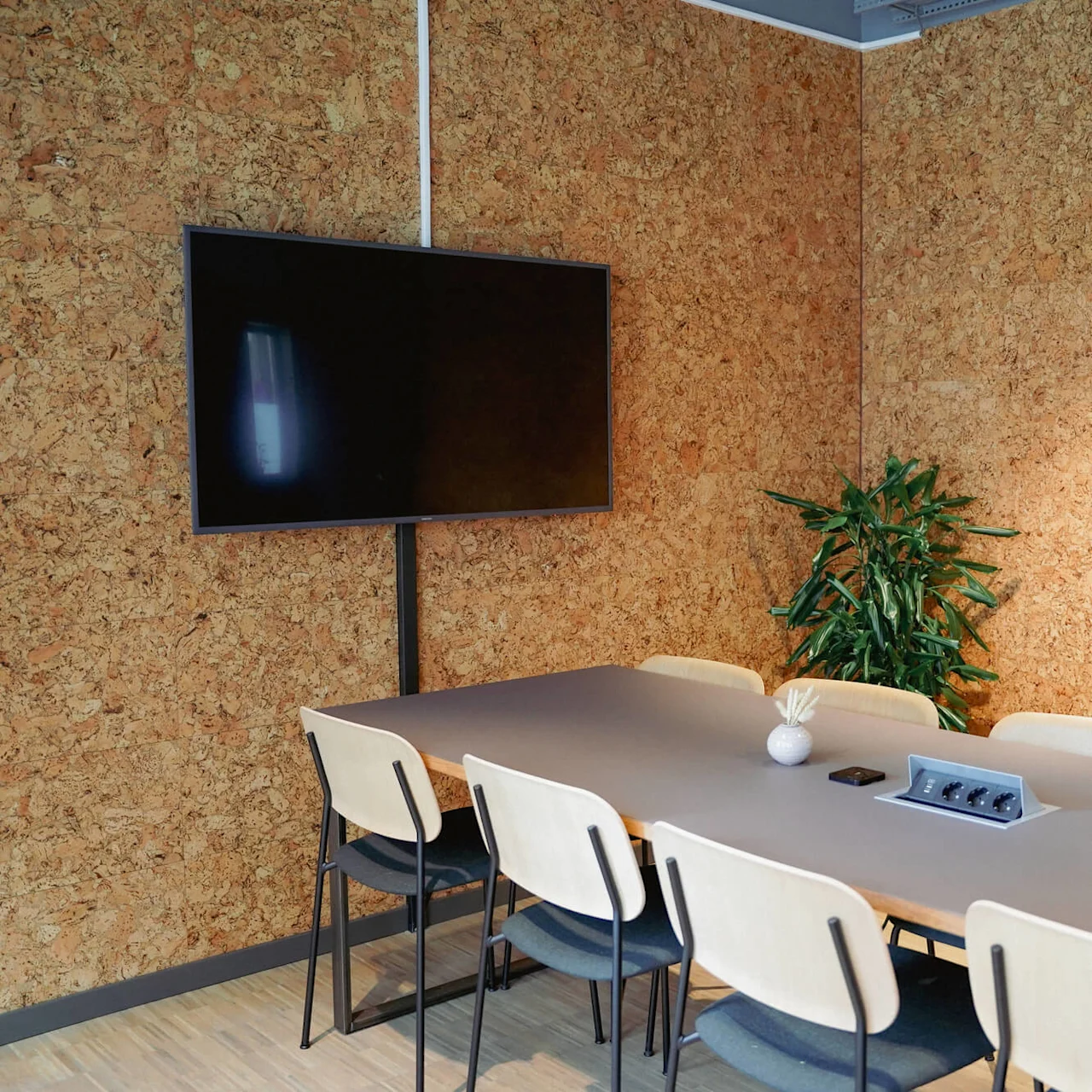 A modern meeting room with a large table, chairs, mounted screen, and a plant, against a cork wall.