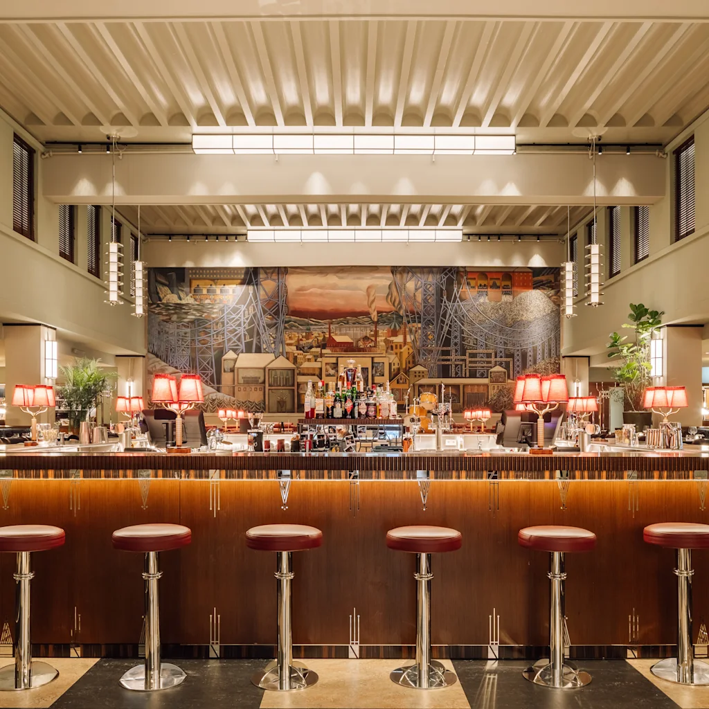 En elegant hotellbar med en lång trädisk, röda pallar och en livlig väggmålning, vilket skapar en varm och inbjudande atmosfär.