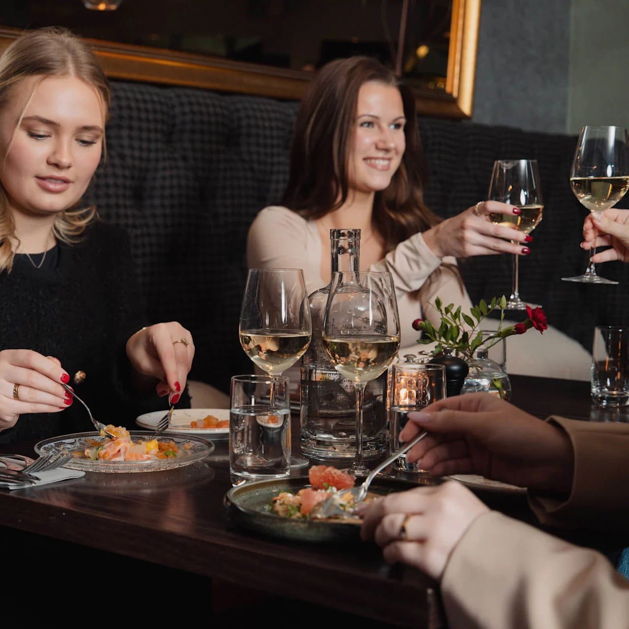 En gruppe mennesker nyder et måltid på en restaurant. En person spiser med bestik, mens en anden løfter et vinglas. Et lys og blomster pynter bordet.