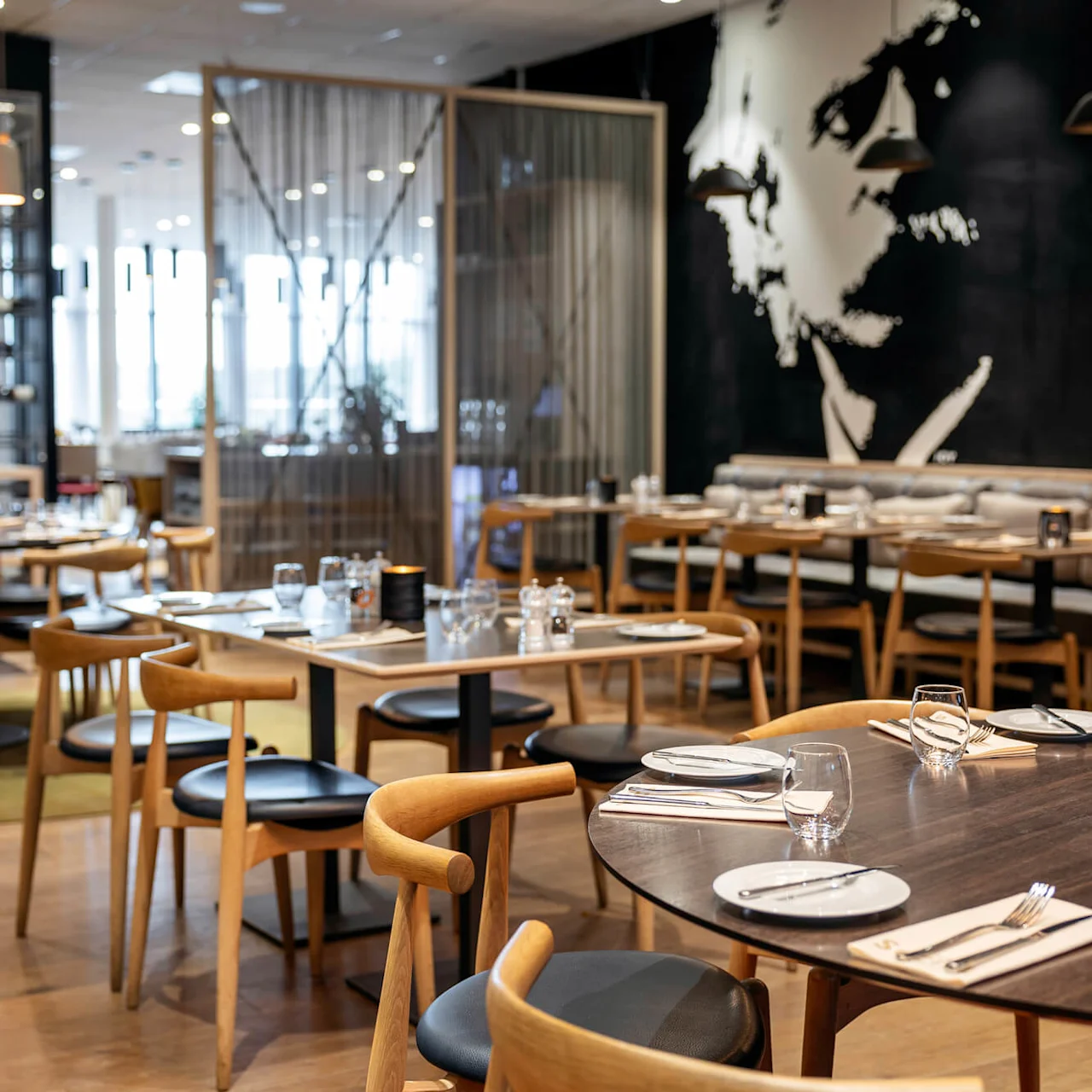 En restaurant med tregulv og oppsatte trebord med tallerkener og glass. Miljøet er moderne med hengelamper og et stort kunstnerisk veggmaleri i bakgrunnen.