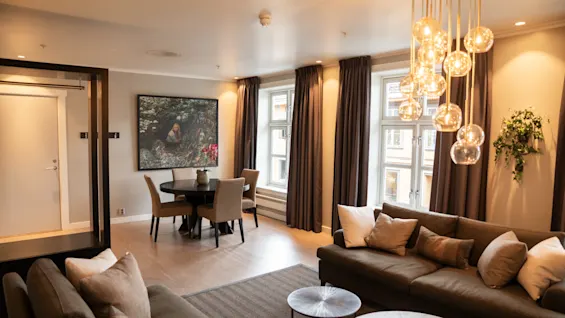 A cozy seating area with modern sofas and cushions sits beneath hanging glass lights in an elegant room featuring large windows, heavy drapes, and a small dining table with chairs.