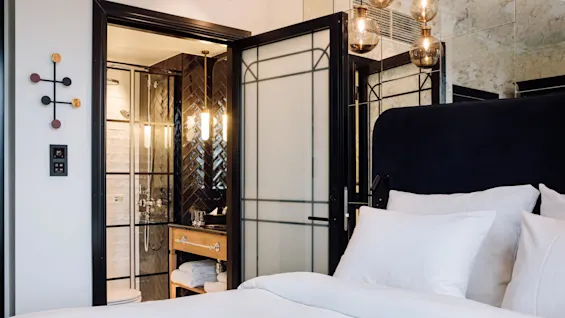 A neatly made bed with white linens in a room adjacent to a modern bathroom with glass doors and wall lighting, featuring a black and wood sink console.