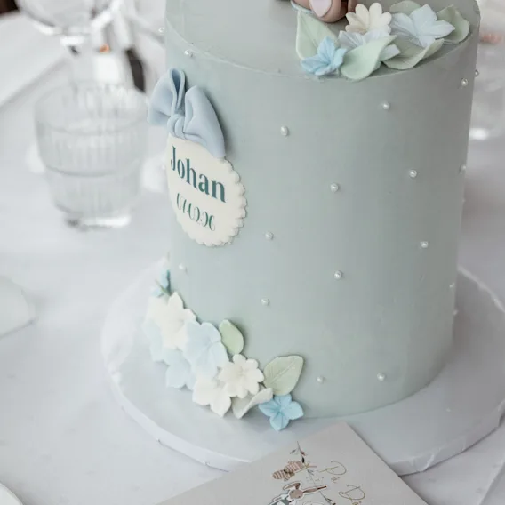 En lyseblå kake dekorert med hvite perler, delikate blomster og et skilt med teksten Johan, som antyder en feiring.