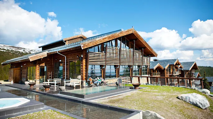 A modern wooden cabin with large glass windows rests in a mountainous landscape. People relax on deck chairs outside, enjoying sunny weather and clear blue skies.